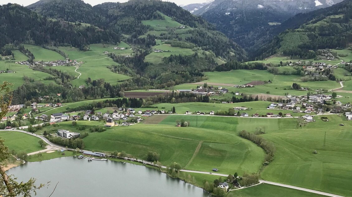 Eine idyllische Landschaft mit sanften Hügeln und einem ruhigen See. Im Hintergrund sind schneebedeckte Berge und kleine Dörfer zu sehen. | © Sabine Schulz
