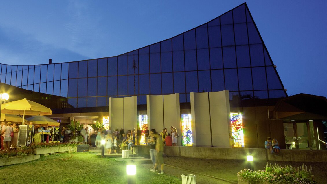 Ein modernes Gebäude mit einer gläsernen Fassade, das bei Dämmerung beleuchtet ist. Vor dem Gebäude befinden sich Menschen und ein grüner Außenbereich. | © Kulturhaus Knittelfeld