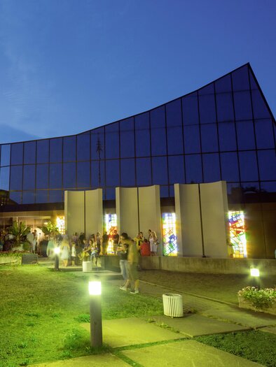 A modern building with a glass facade, illuminated at dusk. In front of the building, there are people and a green outdoor area. | © Kulturhaus Knittelfeld