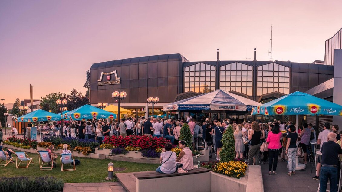 Eine belebte Außenfläche mit vielen Menschen, die sich unter einladenden Markisen versammeln. Der Himmel ist in sanfte Farben getaucht, was eine angenehme Atmosphäre schafft. | © Kulturhaus Knittelfeld