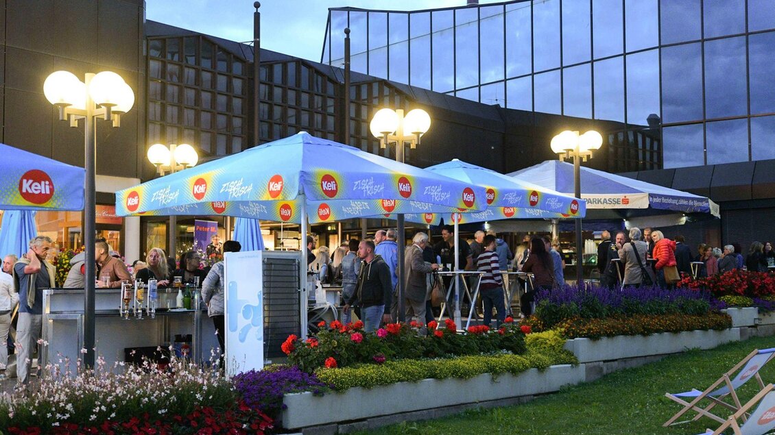 Eine lebhafte Veranstaltung im Freien mit zahlreichen Menschen unter blauen Zelten. Umgeben von bunten Blumen und mit abendlicher Beleuchtung. | © Kulturhaus Knittelfeld