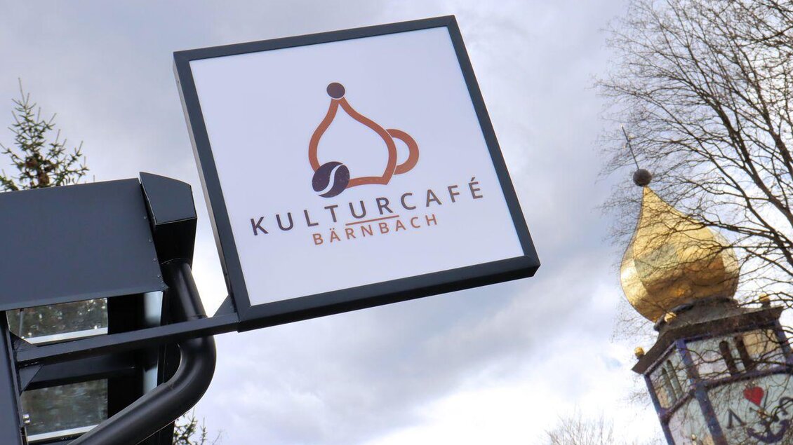 Ein Schild des Kulturcafés in Bärnbach mit einem stilisierten Logo. Im Hintergrund sind Bäume und ein goldener Turm sichtbar. | © Robert Cescutti