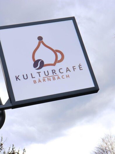 Ein Schild des Kulturcafés in Bärnbach mit einem stilisierten Logo. Im Hintergrund sind Bäume und ein goldener Turm sichtbar. | © Robert Cescutti