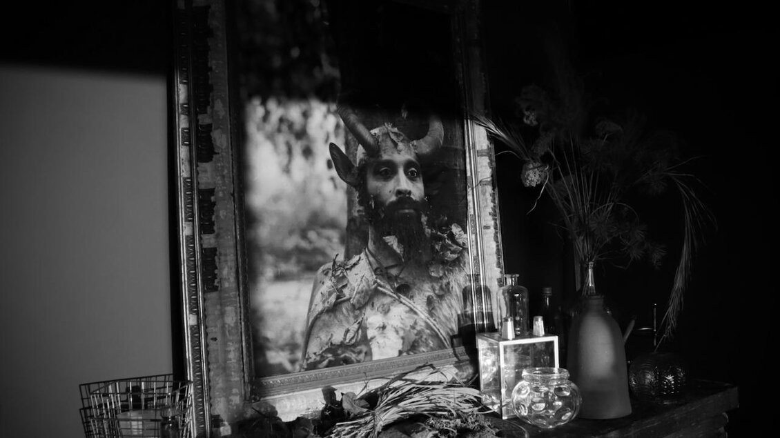 Ein schwarzes und weißes Bild zeigt ein Porträt eines Mannes mit Geweihen. Auf einem Tisch sind verschiedene Gegenstände und Pflanzen verteilt. | © Itshe Petz