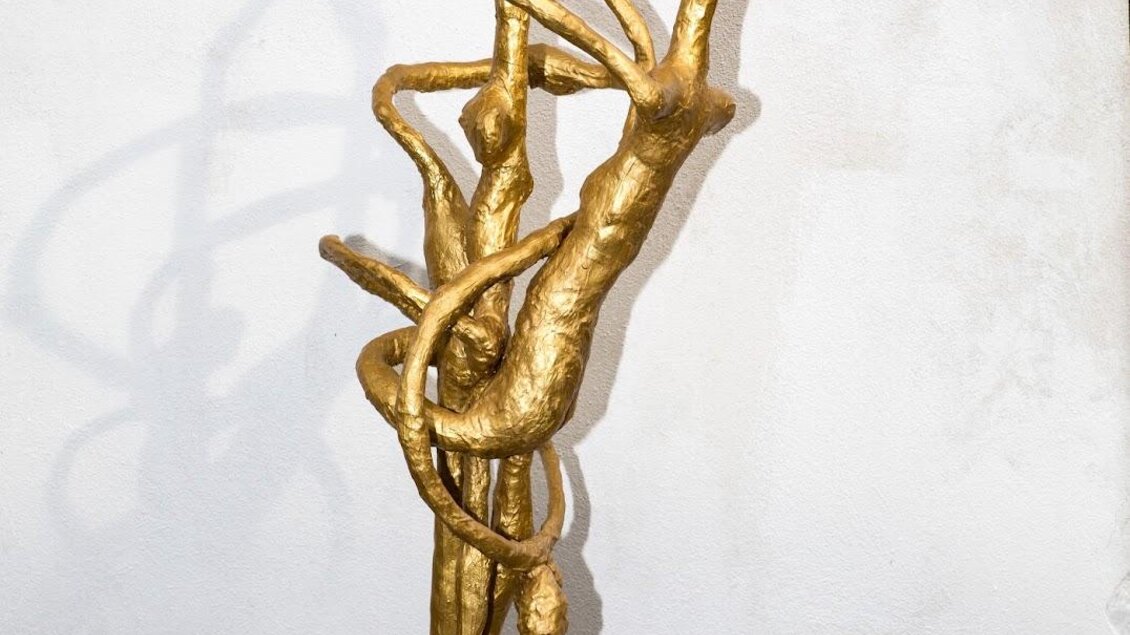 Eine goldene Skulptur, die organic Formen darstellt. Sie zeigt geschwungene Linien und Bewegung auf einem hellen Hintergrund.