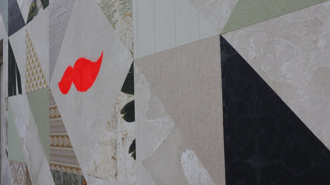 Eine Wand mit geometrischen Mustern in verschiedenen Farben und Texturen. Ein auffälliger roter Kussmund hebt sich von den restlichen Farben ab.