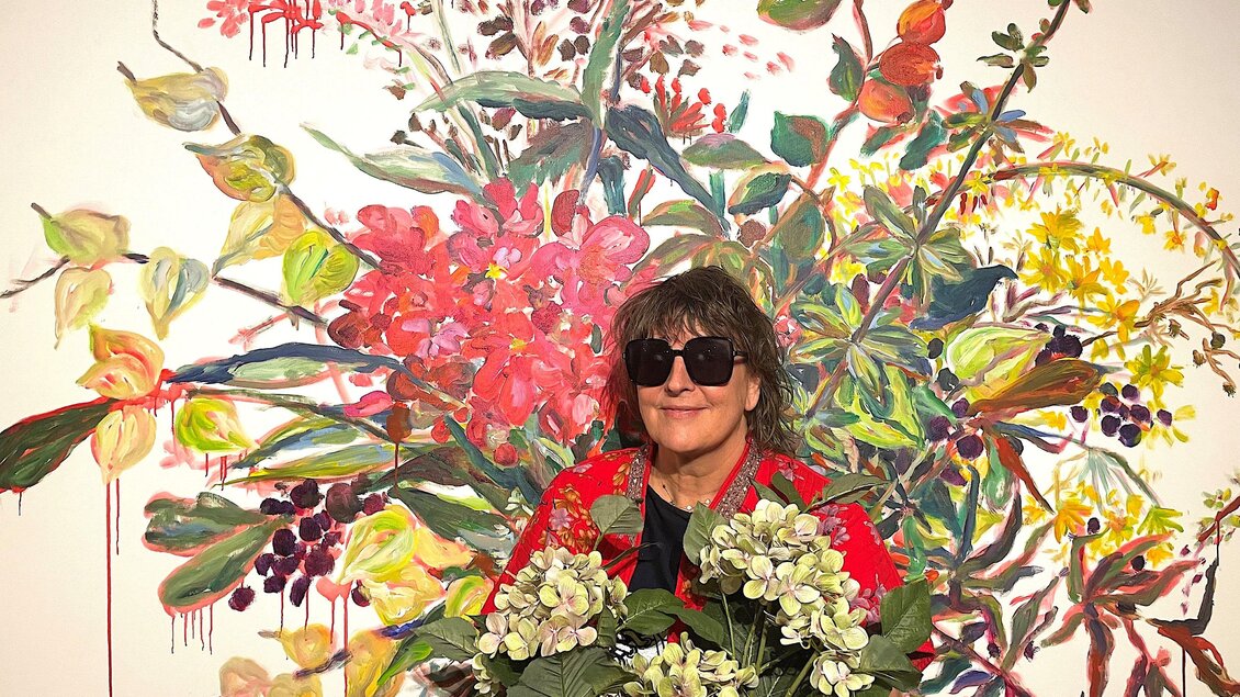 Eine Person mit Sonnenbrille hält einen Strauß Blumen vor einer bunten Blumenkunst. Der Hintergrund zeigt lebendige Farben und florale Motive. | © itshe+io / Kristina Foggensteiner