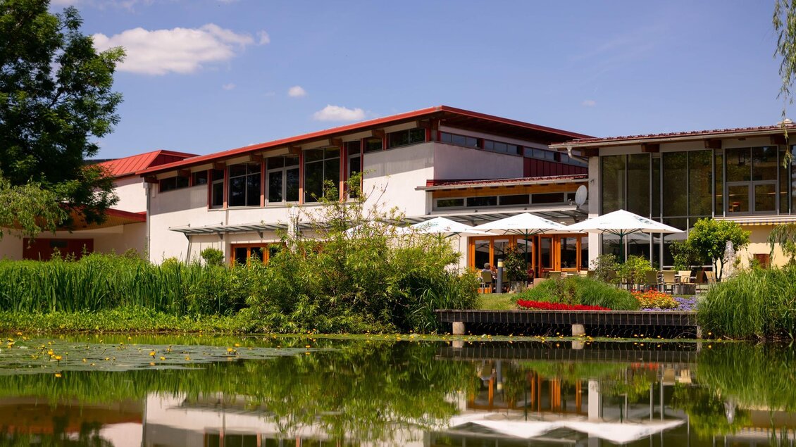 Ein modernes Gebäude mit großen Fenstern und einem roten Dach, umgeben von Grün. Im Vordergrund spiegelt sich der Himmel im ruhigen Wasser eines Teiches. | © TVB Bad Waltersdorf