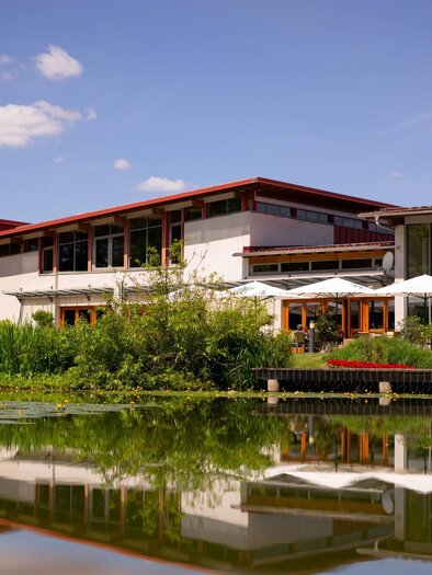 A modern building with large windows and a red roof, surrounded by greenery. In the foreground, the sky is reflected in the tranquil water of a pond. | © TVB Bad Waltersdorf