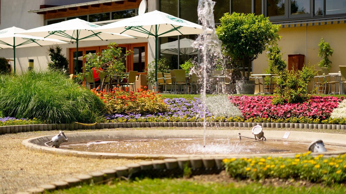 Ein schöner Garten mit einem Brunnen und bunten Blumen. Im Hintergrund sind Tische unter Sonnenschirmen zu sehen. | © Riedenbauer