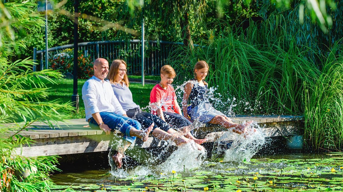 Eine fröhliche Familie sitzt auf einer Holzbank am Wasser und hält ihre Füße im Teich. Spritzendes Wasser und grüne Pflanzen umgeben die Szene. | © Mias Photoart