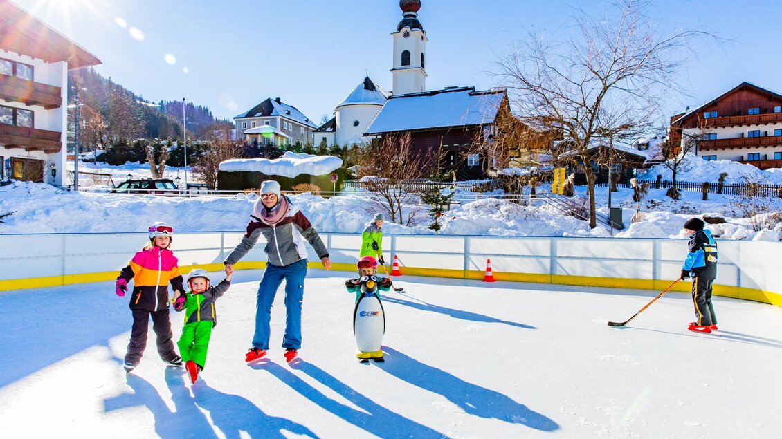 Eine Familie fährt Schlittschuh auf einer Eisbahn inmitten einer schneebedeckten Landschaft. Im Hintergrund sind Häuser und eine Kirche zu sehen. | © Rene Eduard Perhab
