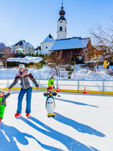 A family is ice skating on a rink in the middle of a snow-covered landscape. In the background, houses and a church can be seen. | © Rene Eduard Perhab