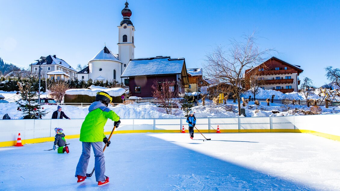 Eine sonnige Eislaufbahn im Winter mit Kindern, die Schlittschuh laufen. Im Hintergrund sind schneebedeckte Häuser und eine Kirche sichtbar. | © Rene Eduard Perhab