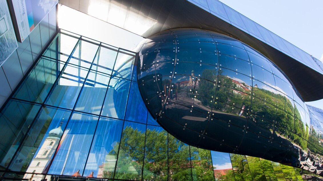 Ein modernes Gebäude mit einer gläsernen Fassade und einer auffälligen, glänzenden Struktur. Die Architektur reflektiert die Umgebung und zeigt einen klaren Himmel im Hintergrund. | © Graz Tourismus - Harry Schiffer