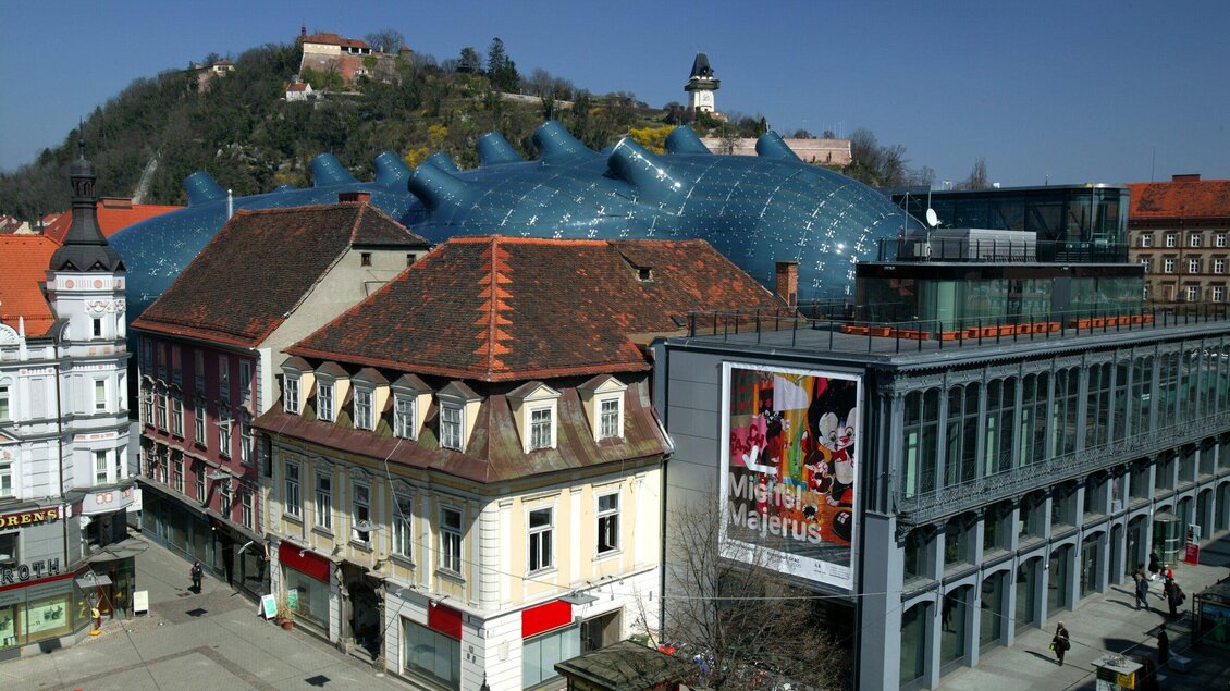 Ein modernes Gebäude mit einer wellenförmigen, blauen Fassade steht neben historischen Häusern. Im Hintergrund ist ein Hügel mit einer Burg zu sehen. | © Graz Tourismus - Harry Schiffer