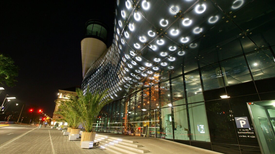 Ein modernes Gebäude mit Glasfassade und faszinierender Beleuchtung in der Nacht. Im Vordergrund befinden sich Pflanzen und ein Parkplatz. | © Graz Tourismus - Harry Schiffer