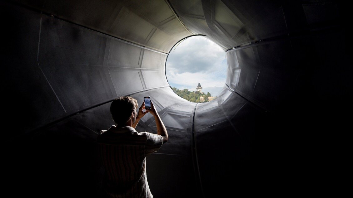 Eine Person steht in einem zylindrischen Raum und fotografiert durch eine runde Öffnung. Im Hintergrund sind Bäume und ein Gebäude vor einem bewölkten Himmel zu sehen. | © Graz Tourismus - Tom Lamm