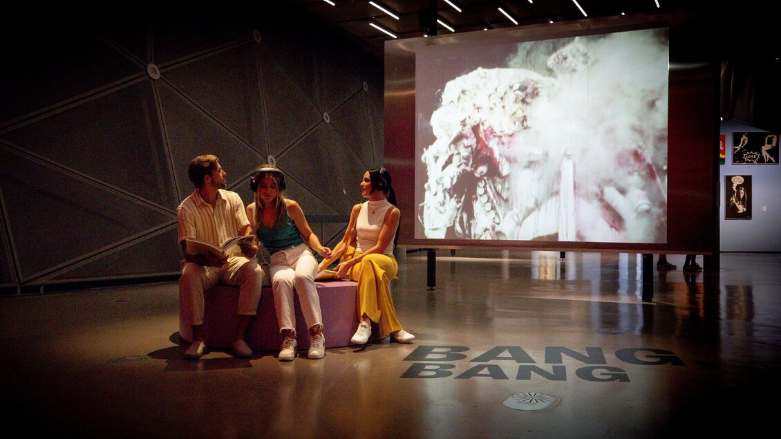 Drei Personen sitzen in einem modernen Ausstellungsraum. Im Hintergrund ist ein großes Bild mit skurrilen Farben und Formen zu sehen. | © Graz Tourismus - Tom Lamm