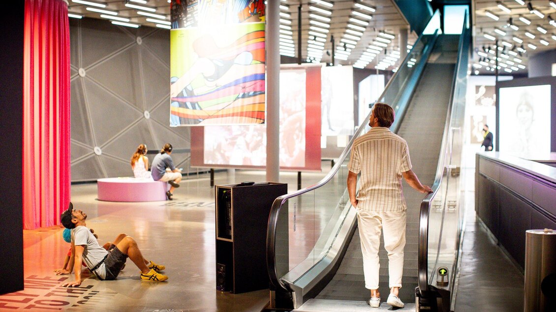 Eine moderne Ausstellungshalle mit einer Rolltreppe. Besucher sitzen entspannt und betrachten die Kunstwerke in der Umgebung. | © Graz Tourismus - Tom Lamm