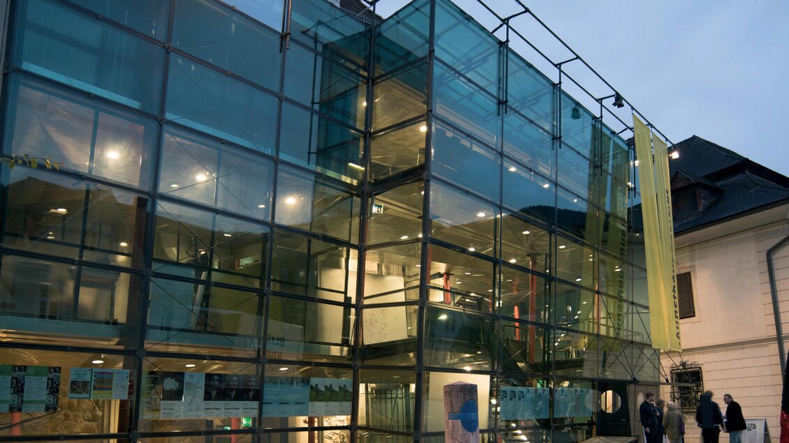 Ein modernes Gebäude mit einer Glasfassade und bunten Elementen. Vor dem Eingang stehen einige Personen und eine Bank ist im Vordergrund sichtbar. | © Flieser Werner