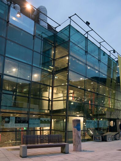 A modern building with a glass facade and colorful elements. In front of the entrance, there are several people, and a bench is visible in the foreground. | © Flieser Werner