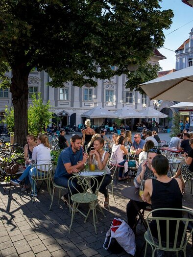 A busy square with many tables outdoors and guests eating and drinking. Bicycles are in the background while the sun is shining. | © Graz Tourismus - Tom Lamm
