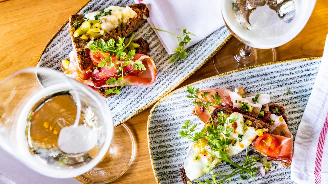 Zwei belegte Brote mit Schinken, Käse und Kräutern, dazu Gläser Sekt auf einem Holztisch. | © Graz Tourismus - Werner Krug