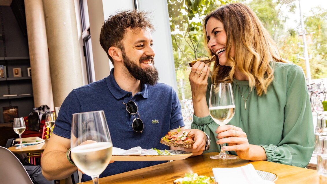 Ein lächelndes Paar genießt ein gemütliches Essen in einem Restaurant. Sie haben Gläser Wasser und eine Platte mit Essen vor sich. | © Graz Tourismus - Werner Krug