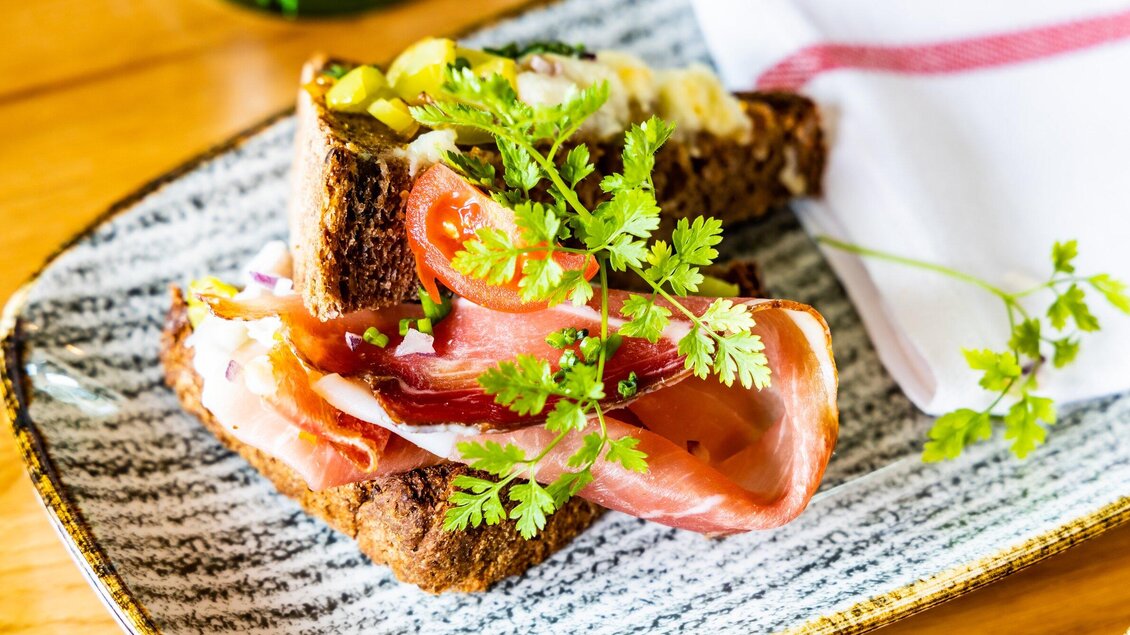 Ein Teller mit zwei Scheiben Brot, belegt mit Schinken, Tomaten und frischen Kräutern. Die Präsentation ist ansprechend und farbenfroh. | © Graz Tourismus - Werner Krug