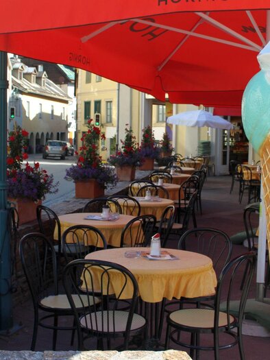 An inviting café with round tables and chairs under red parasols. In the foreground stands a large ice cream cone as decoration. | © TVB Ausseerland - Salzkammergut_Viola Lechner