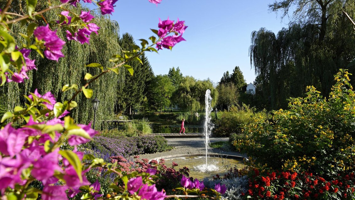 Ein schöner Garten mit bunten Blumen und einem Wasserbrunnen. Im Hintergrund sind Bäume und ein klarer blauer Himmel sichtbar. | © Bernhard Bergmann