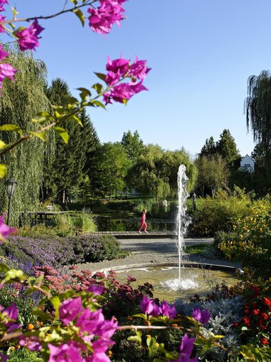 Ein schöner Garten mit bunten Blumen und einem Wasserbrunnen. Im Hintergrund sind Bäume und ein klarer blauer Himmel sichtbar. | © Bernhard Bergmann