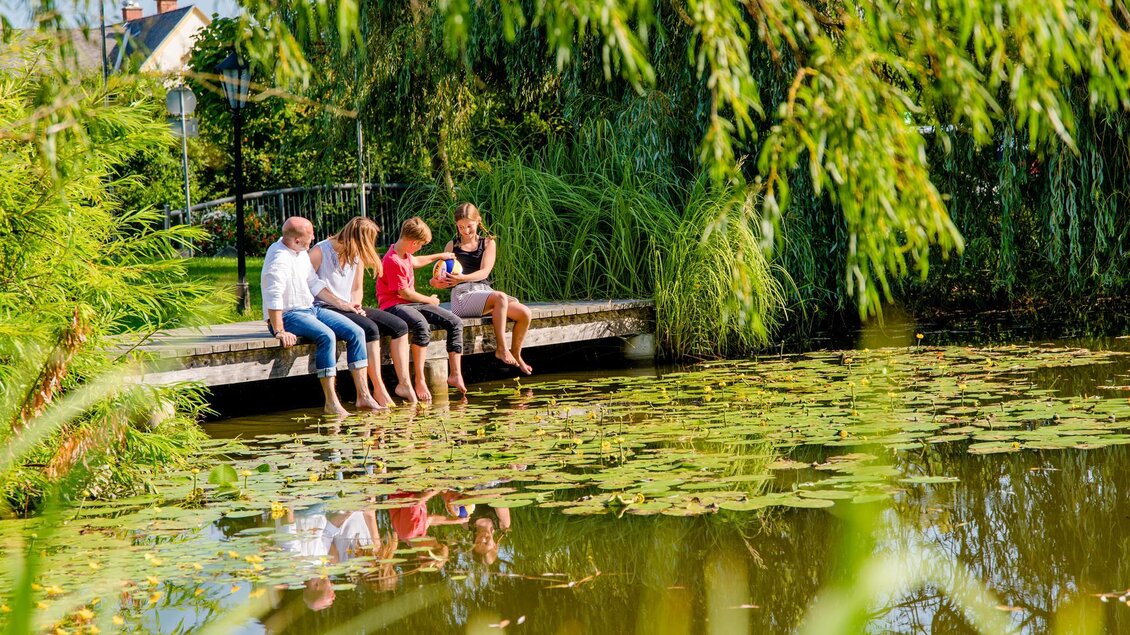 Eine Gruppe von vier Personen sitzt auf einer Holzplanke am Ufer eines ruhigen Teichs. Die Umgebung ist grün und friedlich, mit Schilf und Seerosen im Wasser. | © Mias Photart