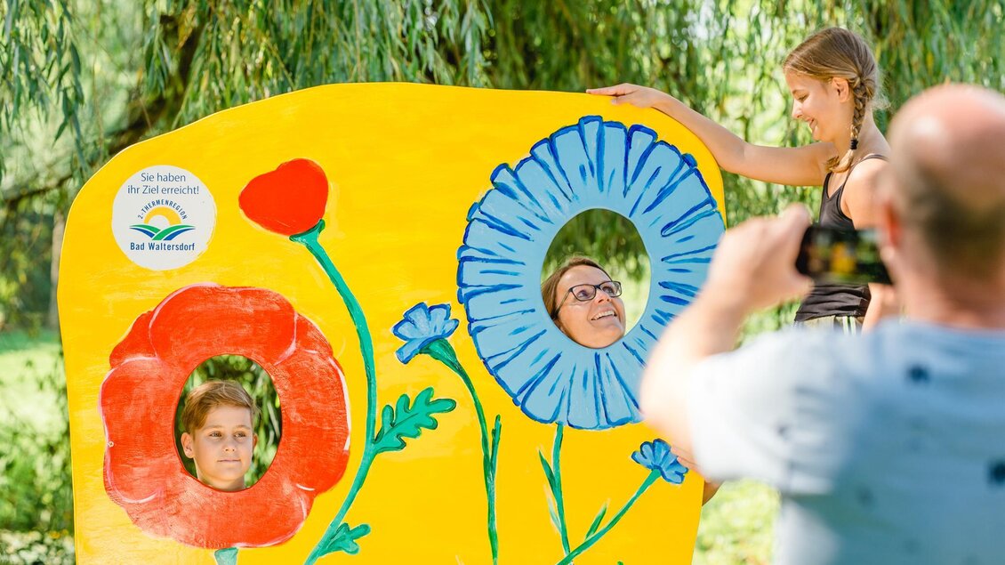 Eine bunte Blütenwand mit Löchern für Gesichter. Kinder und Erwachsene posieren und machen Fotos davor. | © Mias Photoart