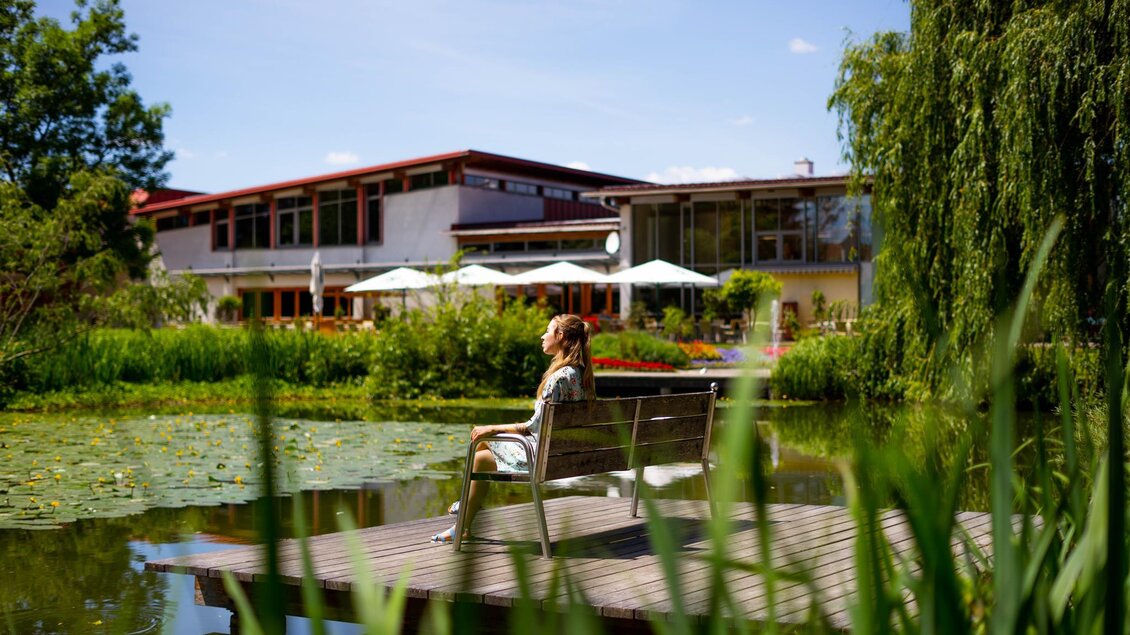 Eine ruhige Szene an einem Teich mit einer Person, die auf einer Bank sitzt. Im Hintergrund ist ein modern gebäudeter Raum und viel Grün zu sehen.