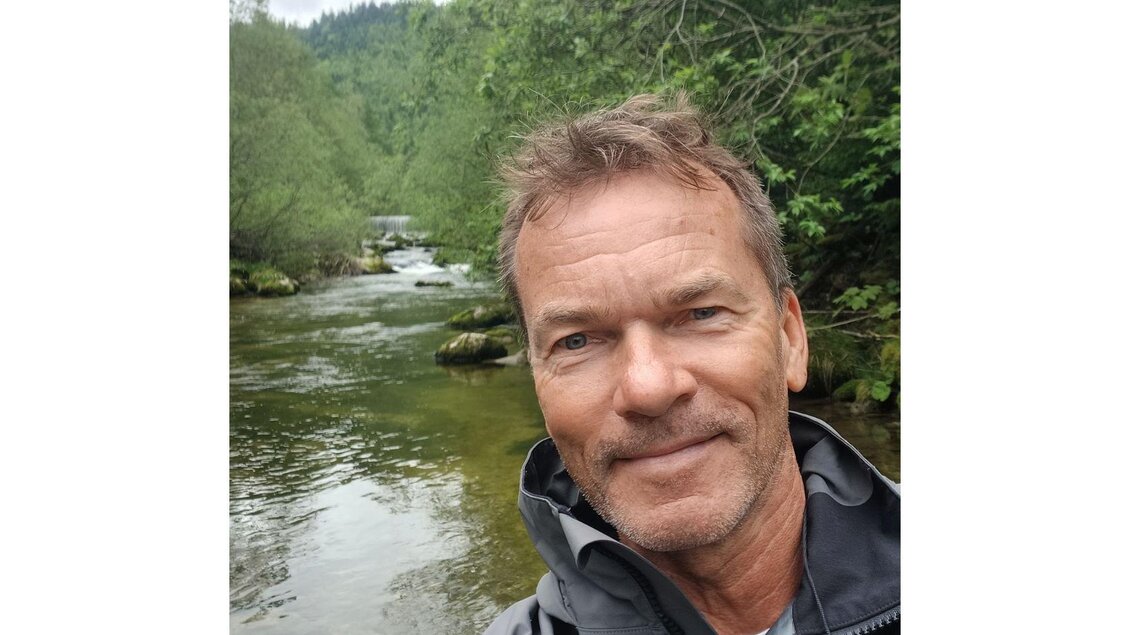 Ein Mann steht am Ufer eines ruhigen Flusses, umgeben von üppigem Grün. Die natürliche Landschaft ist friedlich und einladend. | © Kurt Weber