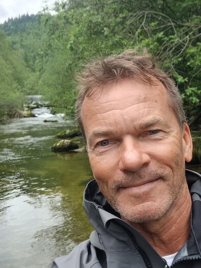 A man stands at the shore of a calm river, surrounded by lush greenery. The natural landscape is peaceful and inviting. | © Kurt Weber