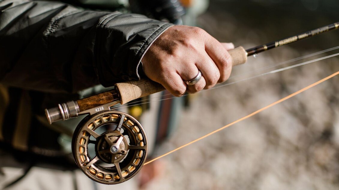 Ein Fischer hält eine Angelrute in der Hand. Die Spule der Rute ist gut zu erkennen und bereit zum Angeln. | © TVB Ausseerland Salzkammergut_Katrin Kerschbaumer