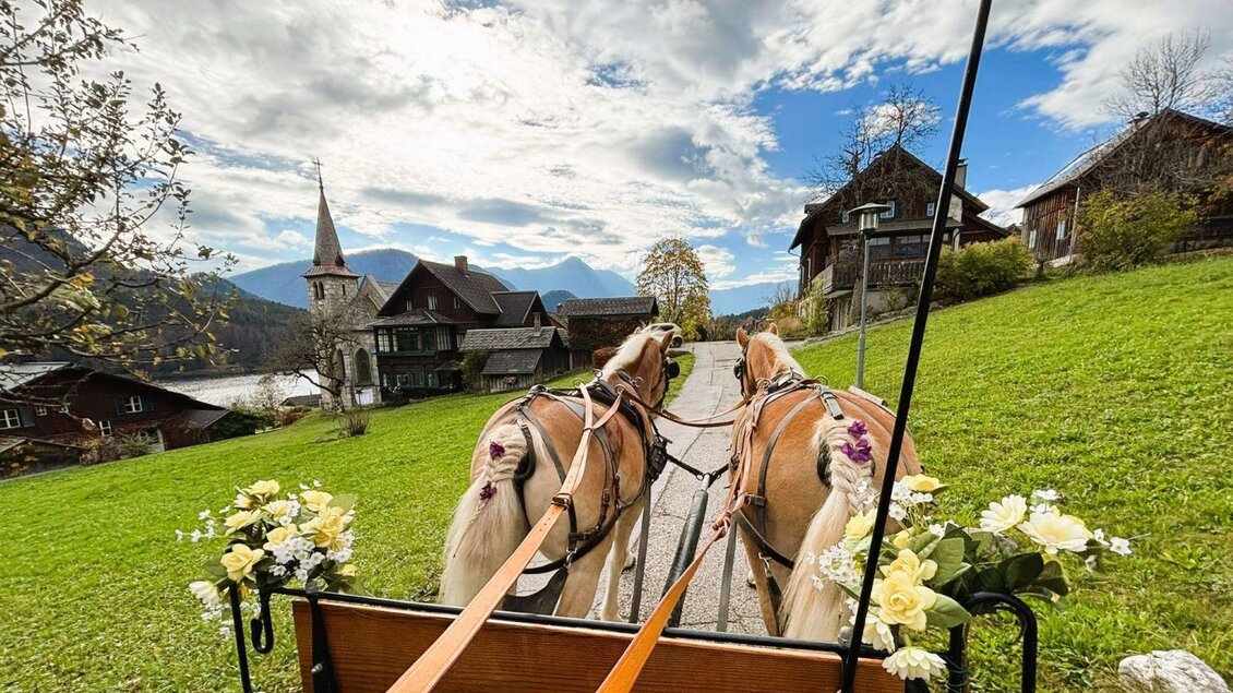 Eine Pferdekutsche mit zwei Pferden fährt durch eine malerische Landschaft. Im Hintergrund sind grüne Wiesen und traditionelle Holzhäuser sowie ein Kirchturm zu sehen. | © www.pferdefreunde-zloam.at