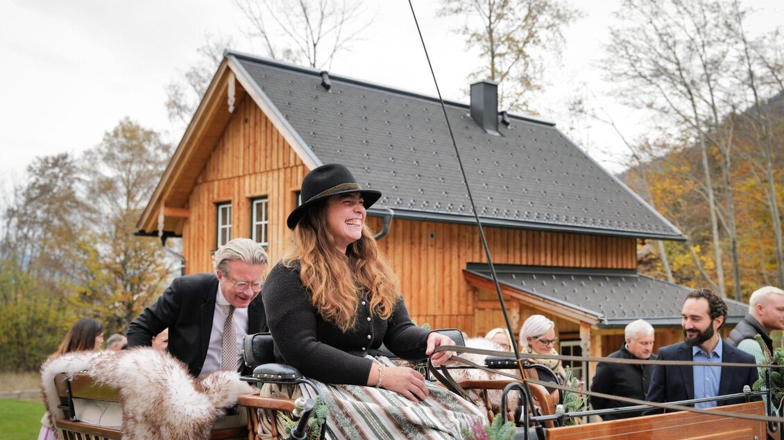 Eine fröhliche Gesellschaft fährt mit einer Kutsche vor einem Holzhaus. Die junge Frau trägt einen Hut und lächelt, während die anderen Gäste im Hintergrund stehen. | © Narzissendorf Zloam