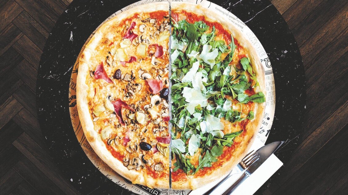 Eine halbierte Pizza mit einer Seite belegt mit Wurst, Pilzen und Oliven und der anderen Seite mit frischem Rucola und Parmesan. Die Pizza liegt auf einem dunklen Tisch mit Besteck daneben. | © L'Osteria