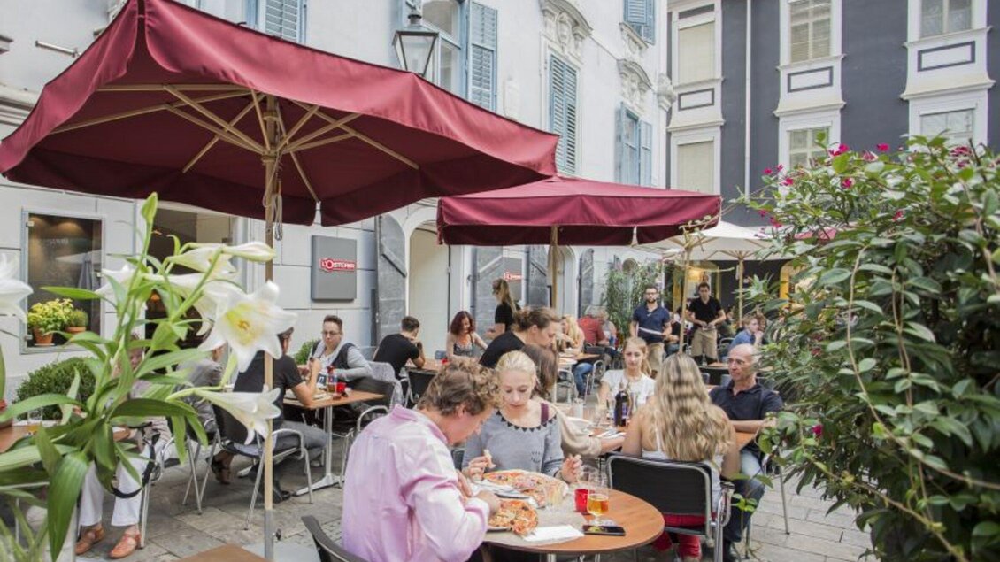 Ein gemütliches Restaurant mit roten Sonnenschirmen im Freien. Viele Gäste genießen ihr Essen und die entspannte Atmosphäre. | © L'Osteria