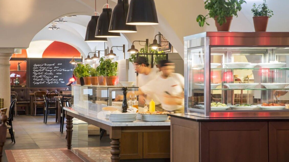 Ein einladendes Restaurant mit einer offenen Theke und einer gemütlichen Atmosphäre. Köche bereiten Gerichte vor, während Pflanzen das Ambiente auflockern. | © L'Osteria