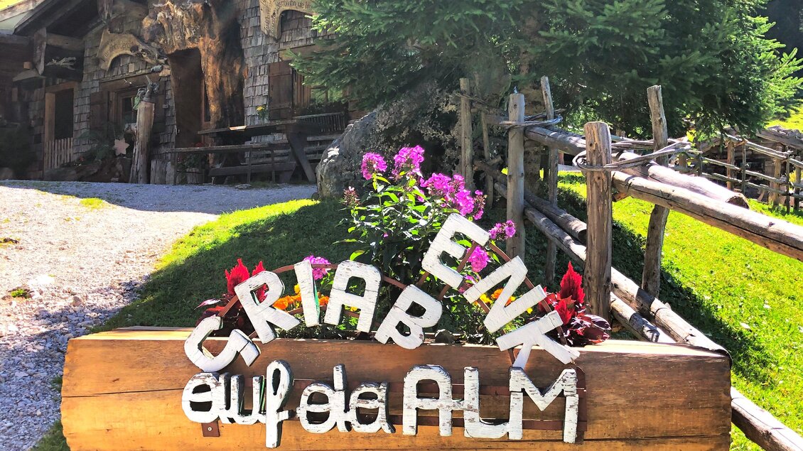 Ein Holzschild mit bunten Buchstaben steht auf einer Wiese. Im Hintergrund sind Bäume und ein traditionelles Gebäude zu sehen. | © ©KajetanGerharter 