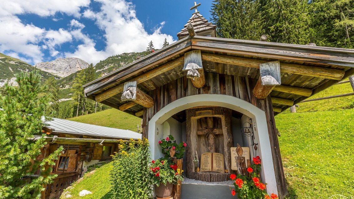 Ein charmantes Holzhaus umgeben von grüner Landschaft und Bergen. Bunte Blumen schmücken den Eingangsbereich. | © ©KajetanGerharter 
