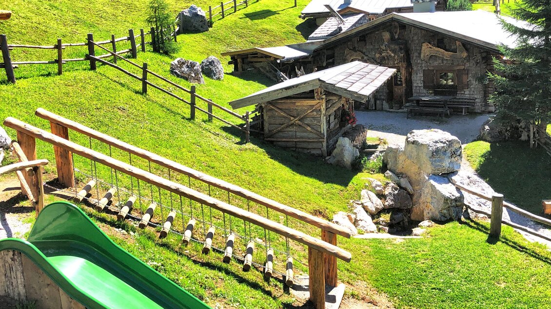 Ein Spielplatz in der Natur mit einer grünen Rutsche und einem kleinen Holzgebäude im Hintergrund. Umgeben von grünen Wiesen und Bäumen. | © ©KajetanGerharter 