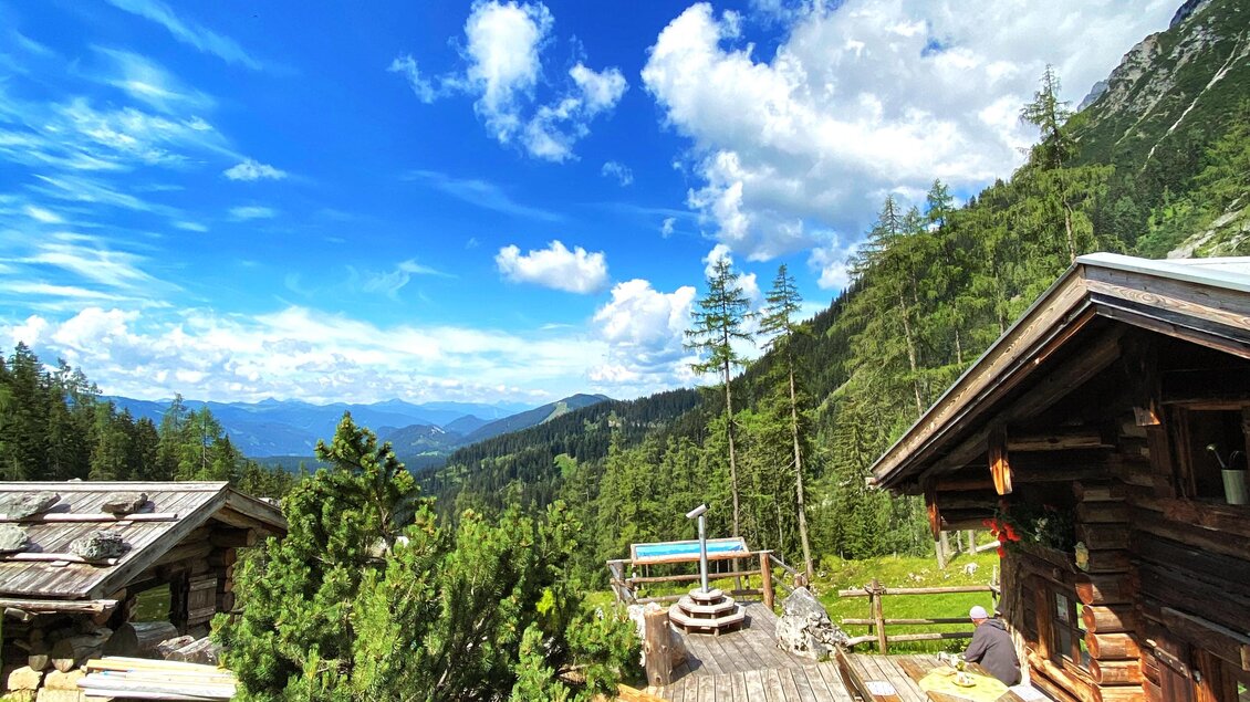 Ein gemütliches Holzhaus umgeben von grünen Bäumen und Bergen. Der Himmel ist blau mit einigen Wolken, und eine Holzterrasse lädt zum Entspannen ein. | © ©KajetanGerharter 