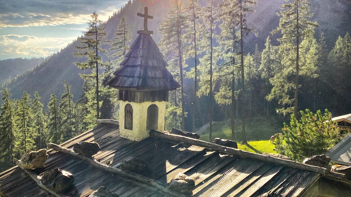 Eine kleine Kapelle in den Bergen, umgeben von hohen Bäumen. Die Sonne scheint durch die Wolken und beleuchtet die Szene. | © ©KajetanGerharter 