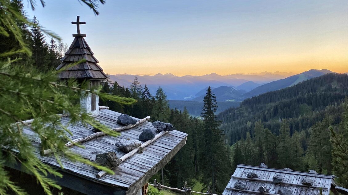 Eine malerische Berglandschaft mit einer kleinen Kapelle und einem Holzhaus. Der Sonnenuntergang verleiht dem Himmel eine warme Farbe über den Bergen. | © ©KajetanGerharter 